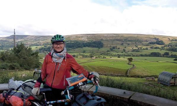 A woman is standing with a fully loaded touring bike. She is wearing a red winter cycling jacket, scarf and green helmet. Behind her is a lot of country scenery