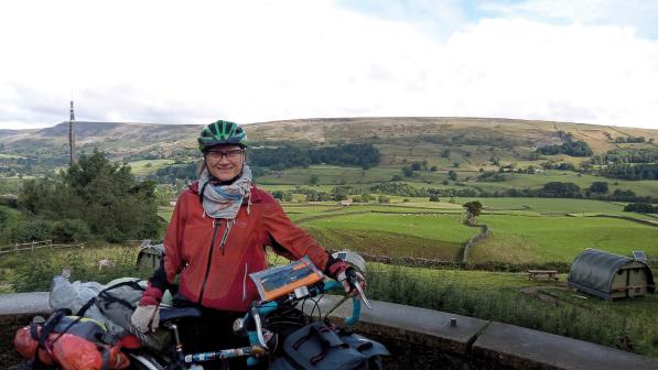 A woman is standing with a fully loaded touring bike. She is wearing a red winter cycling jacket, scarf and green helmet. Behind her is a lot of country scenery