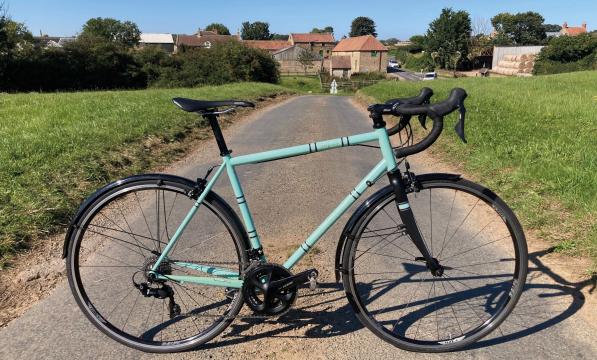 A pale turquoise road bike with mudguards propped up on a narrow country lane