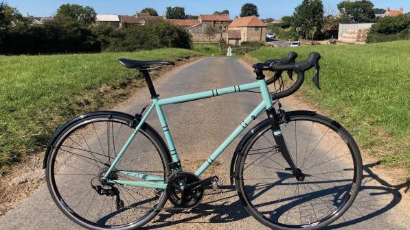 A pale turquoise road bike with mudguards propped up on a narrow country lane