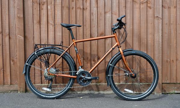 A copper-coloured flat-bar touring bike with black rear rack and mudguards leaning against a wooden fence