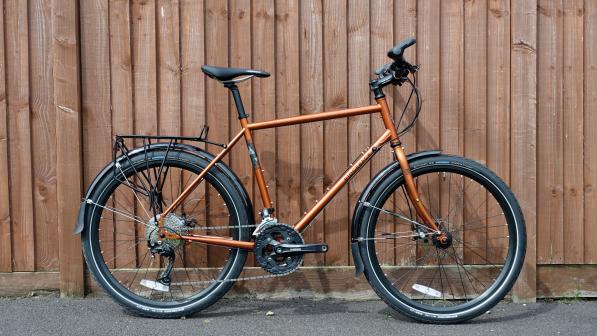A copper-coloured flat-bar touring bike with black rear rack and mudguards leaning against a wooden fence