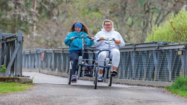 Two women are cycling a side-by-side tandem trike across a bridge. They are wearing tracksuits and are smiling