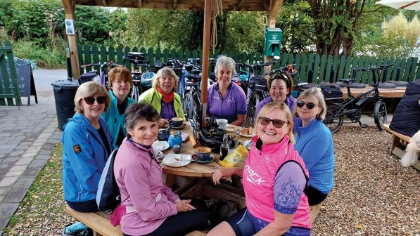 A group of women are sitting around a picnic table at a cafe. There's a selection of drinks and snacks in front of them. They're wearing cycling kit and their bikes can be seen leaning against a fence in the background. They are smiling at the camera.