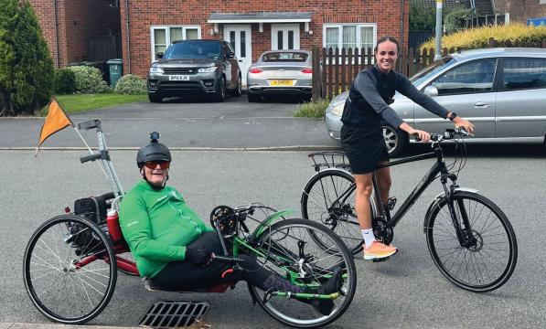A man on a handcycle with a woman on a hybrid bike. He's wearing a green cycling jersey and black leggings. She's wearing black cycling gear and trainers.
