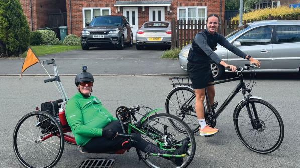 A man on a handcycle with a woman on a hybrid bike. He's wearing a green cycling jersey and black leggings. She's wearing black cycling gear and trainers.