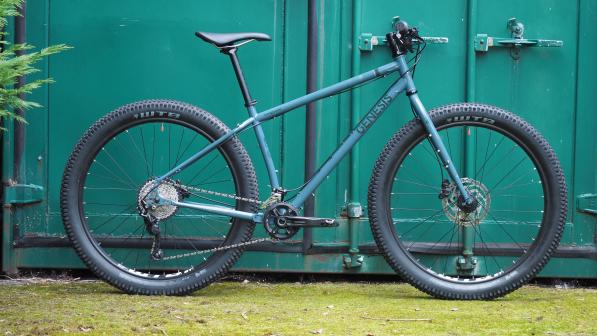 A rigid mountain bike with fat tyres leaning against a turquoise lock-up, The bike is blue and has the Genesis logo on it