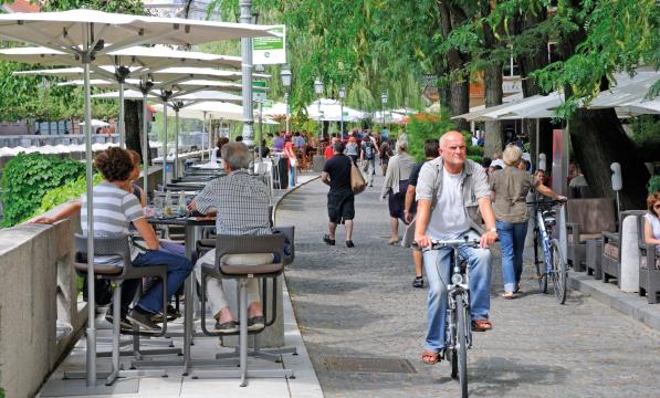 A man is cycling along a shared cycle and walking path next with restaurants on one side and people sitting at tables eating and drinking on the other