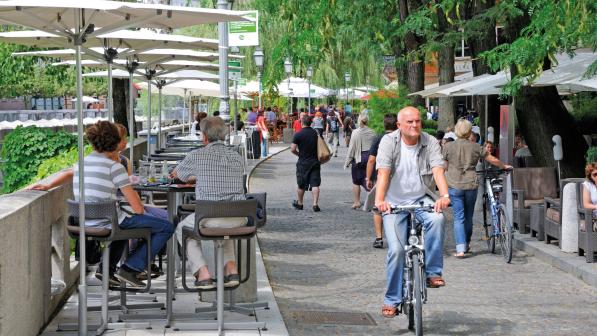 A man is cycling along a shared cycle and walking path next with restaurants on one side and people sitting at tables eating and drinking on the other