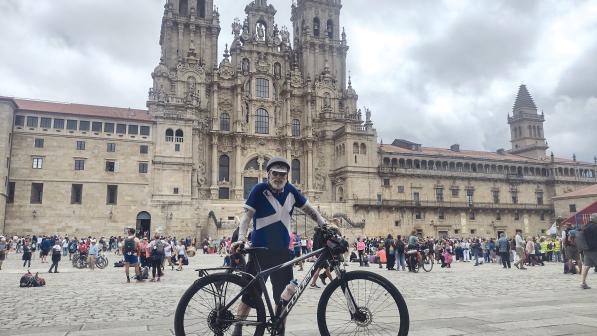 A man in a blue and white cross of St Andrew jersey, black shorts, cycling helmet and sunglasses is standing with a loaded touring bike in front of a cathedral
