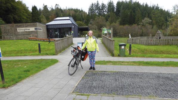A woman is standing next to her packed touring bike in a park. She is wearing patterned leggings and a hi-vis yellow jacket.