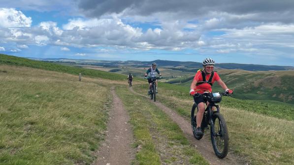 Three women are cycling on mountain bikes along an off-road track through hills in Northumberland. They are wearing MTB clothing, cycling sunglasses and helmets.