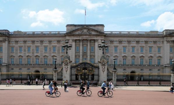 Three people on Santander hire bikes are riding past the gates of Buckingham Palace in London. There are people walking around in the background.