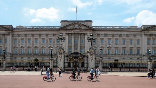 Three people on Santander hire bikes are riding past the gates of Buckingham Palace in London. There are people walking around in the background.