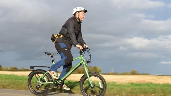 A man is riding a green e-bike with 20in wheels. He's wearing jeans, trainers, a jacket and cycling helmet. He's standing out of the saddle.