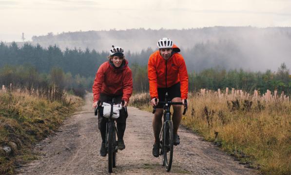Two people are cycling along an off-road track through mountainous countryside. They are on loaded gravel bikes and wearing waterproof cycling kit