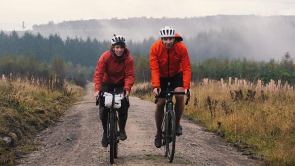 Two people are cycling along an off-road track through mountainous countryside. They are on loaded gravel bikes and wearing waterproof cycling kit