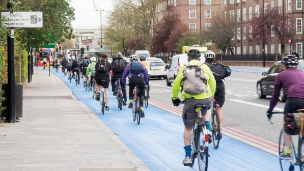Lots of cyclists are riding along a blue segregated cycle lane next to a busy urban road. They are on a variety of bikes and wearing all kinds of clothing. Several have backpacks and it looks like they're cycling to work