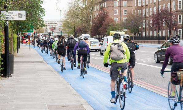 Lots of cyclists are riding along a blue segregated cycle lane next to a busy urban road. They are on a variety of bikes and wearing all kinds of clothing. Several have backpacks and it looks like they're cycling to work