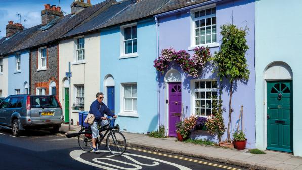A woman wearing blue and white striped three-quarter length trousers and a blue top is cycling over a 20 MPH logo painted on a road. She is going past a row of pastel painted houses. She is wearing sunglasses and has a shoulder bag across her body