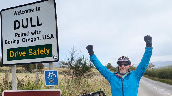 Mark Wedgewood standing next to the sign for Dull Scotland with his arms in the air and his bike leaning beside him