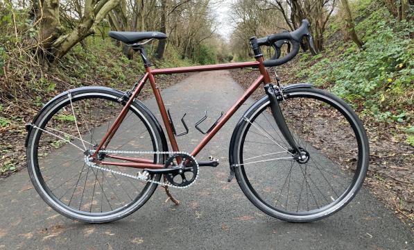 The Spa Cycles Audax Mono, a bronze fixed-wheel bike, propped up on a tarmac tree-lined path 