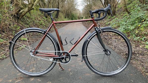 The Spa Cycles Audax Mono, a bronze fixed-wheel bike, propped up on a tarmac tree-lined path 