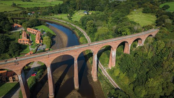 An aerial shot of a Victorian viaduct over a river. It disappears into some trees. A railway line runs under it and there are houses and cars in the background