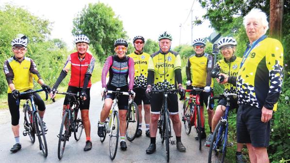 A mixed group of people are all standing with their bikes. They are mostly wearing yellow and black club kit. A couple of people are in non-club cycling kit. They are wearing helmets. They are all smiling at the camera