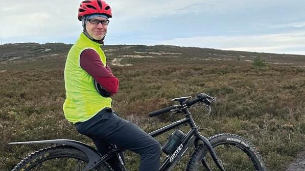 A man is sitting astride a mountain bike on a muddy path. He's got his arms crossed and is smiling at the camera. He's wearing MTB trousers, a red helmet and jersey and a bright yellow gilet.