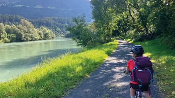 A child is cycling along a gravel track alongside a river. There are mountains in the background and the track is lined with trees. The child is on a mountain bike and wearing shorts and T-shirt, with a cycle helmet and backpack