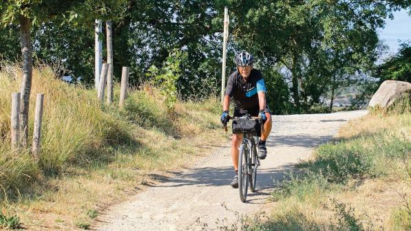 A man is riding along a gravel track through countryside. He's riding a touring bike with a handlebar bag on the front. He's wearing blue and black cycling short-sleeved jersey and shorts, a cycle helmet and glasses