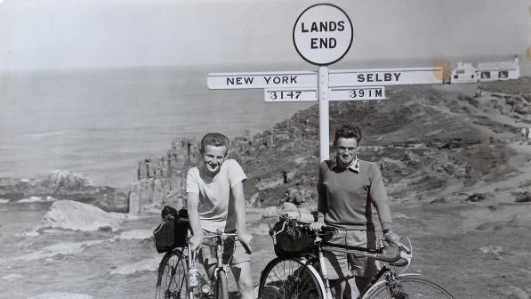 A scan of an old black and white photo. Two men are standing underneath the Land's End sign with their bikes. They are wearing normal clothes and shoes: shorts and a T-shirt and shorts, shirt and sweater. In the background you can see the sea and a cliff. The sign points left and reads New York with 3147 underneath and right and reads Selby with 391 underneath.