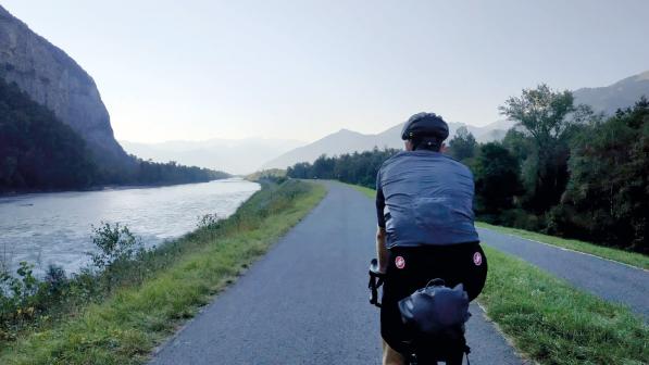 A man is cycling away from the camera on a paved cycle path alongside a river, with mountains in the background. He's wearing cycling kit and riding a touring bike with a packed seatpack