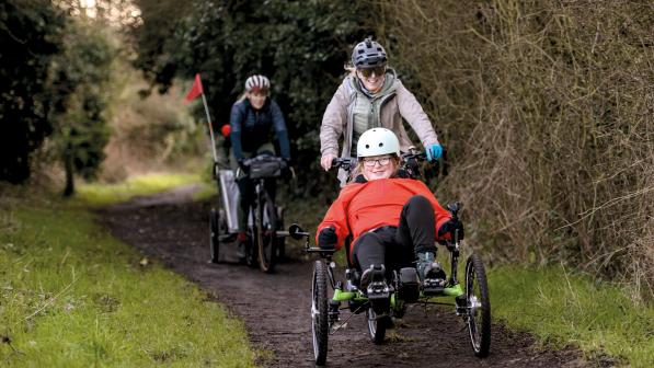 Three people are cycling along a muddy off-road track. A girl on recumbent trike is in front, she is wearing a bright orange jacket. Behind her is a woman on flatbar bike and behind her is a woman on a gravel bike with a trailer on the back