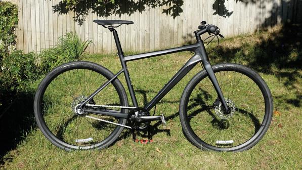 The Boardman URB 8.6 commuter bike, a dark grey hybrid bike with a flat handlebar, propped up in a garden with a wooden fence in the background