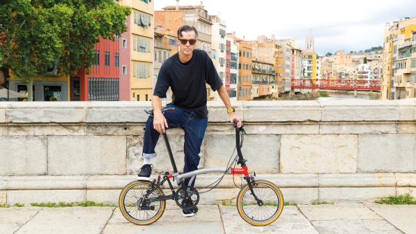 A man in blue jeans and black T-shirt is sitting on the wall of a bridge over a canal in a city. He is holding an unfolded Brompton bike in front of him