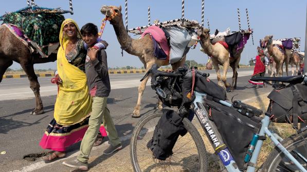 A woman in a sari and a young boy are walking past a loaded touring bike and smiling. Behind them is a line of camels with saddlebags and tables on their backs