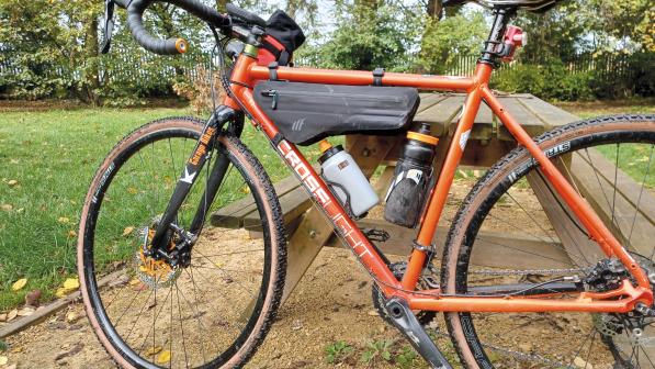 An orange gravel bike is leaning against a wooden table outside. The Wedge Frame Bag is attached to the top tube with water bottles in cages on the down tube and seat tube