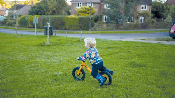 A young boy is riding a yellow Frog balance bike on a green. He's wearing a patterned sweater with buses on it and patterned wellies with dinosaurs on them