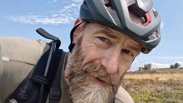 A man has taken a selfie riding. He's wearing a helmet, he's got a fine beard. There's countryside in the background