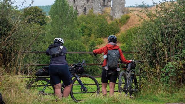 Two people with rucksacks and mountain bikes are looking over a metal gate at a ruined castle