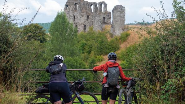 Two people with rucksacks and mountain bikes are looking over a metal gate at a ruined castle