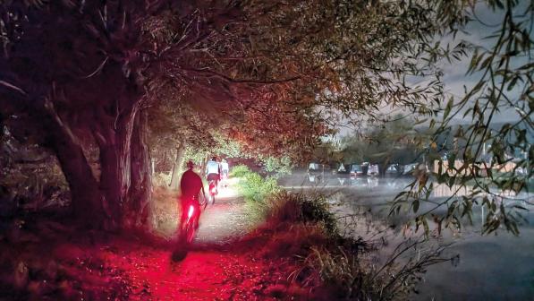 A night-time shot of cyclists riding along a canal towpath from behind. The cyclists' rear lights are shining red in the trees and there are narrow boats on the canal