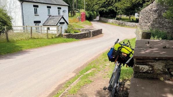 A loaded touring bike is leaning against a stone wall in a village with a white-washed house and a stone-walled house in the background