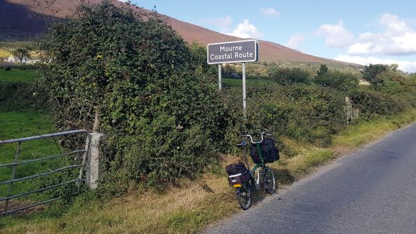 A Brompton bike with front and back bags is leaning against a road sign on a country road. The sign reads 'Mourne Coastal Route'
