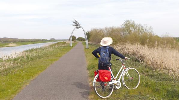 A person is standing on a grass verge next to a paved path looking out over fields. They are holding a white hybrid bike with panniers and wearing black clothes, a white sun hat and blue rucksack