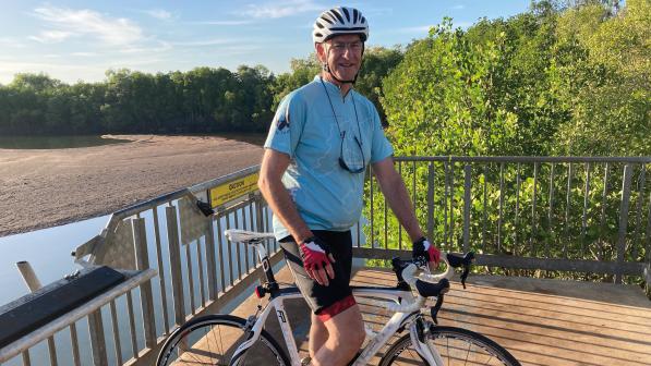 A man is standing on a wooden deck astride a white road bike. He's wearing a cycling helmet and kit. Behind the deck is a water and some trees