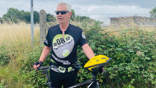 A man wearing cycling kit and sunglasses is standing holding his bike. His yellow AA cycling helmet is on the bike handlebar. He's in front of a hedge