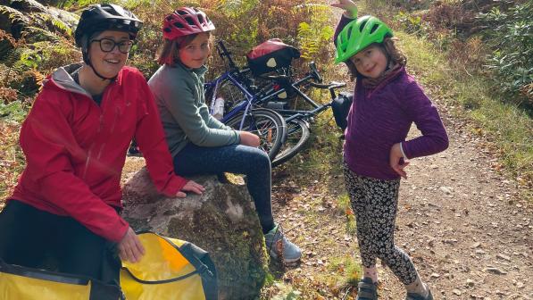 Three people (a woman and two children) are next to an off-road trail. They are all wearing sports gear and cycling helmets. Two kids bikes can be seen in the background and in the foreground are packed panniers